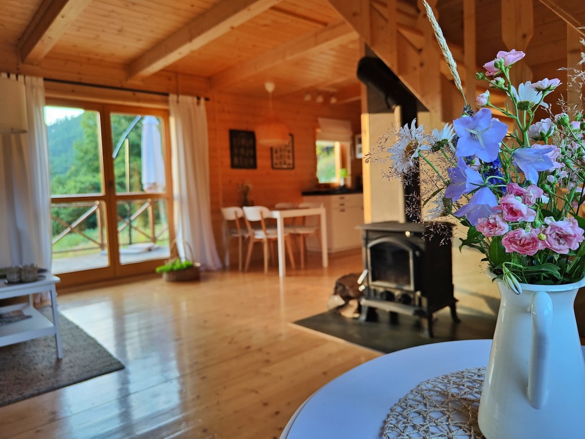 A cozy interior includes a wooden ceiling and large windows that invite natural light. A stove is positioned near a seating area, and a vase with flowers adds a touch of color to the table. Surrounding greenery offers a peaceful view.