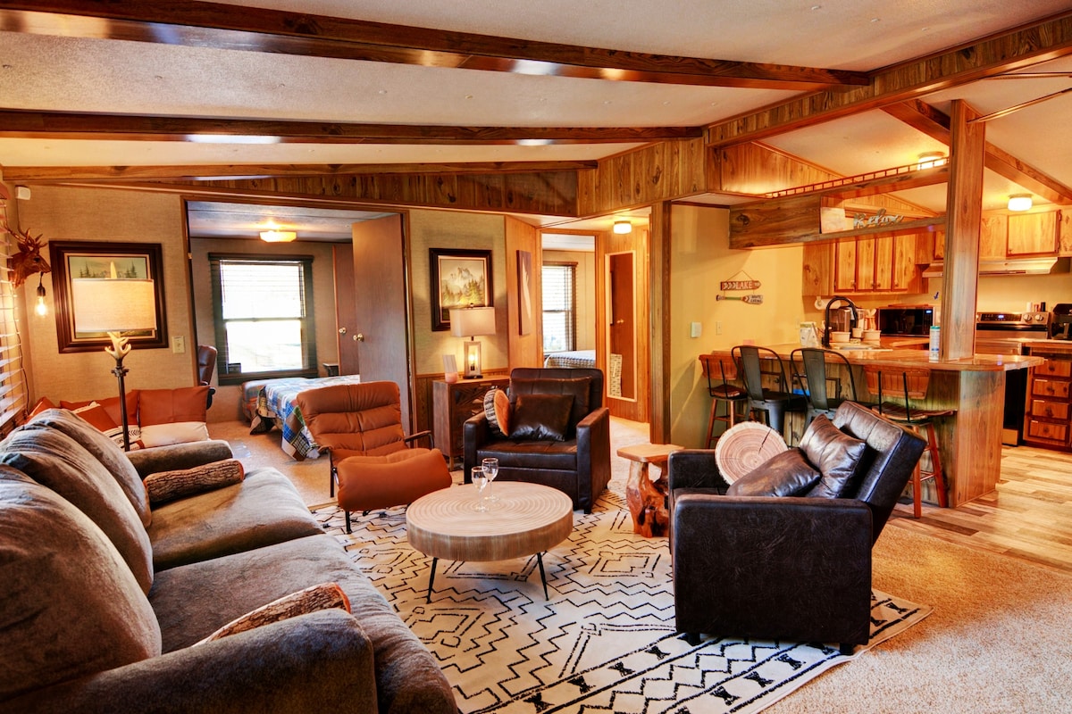 A spacious living area features comfortable seating, including a sectional sofa and two armchairs positioned around a round coffee table. Warm wood accents and visible textures enhance the inviting style. The open layout connects seamlessly to the kitchen area in the background.