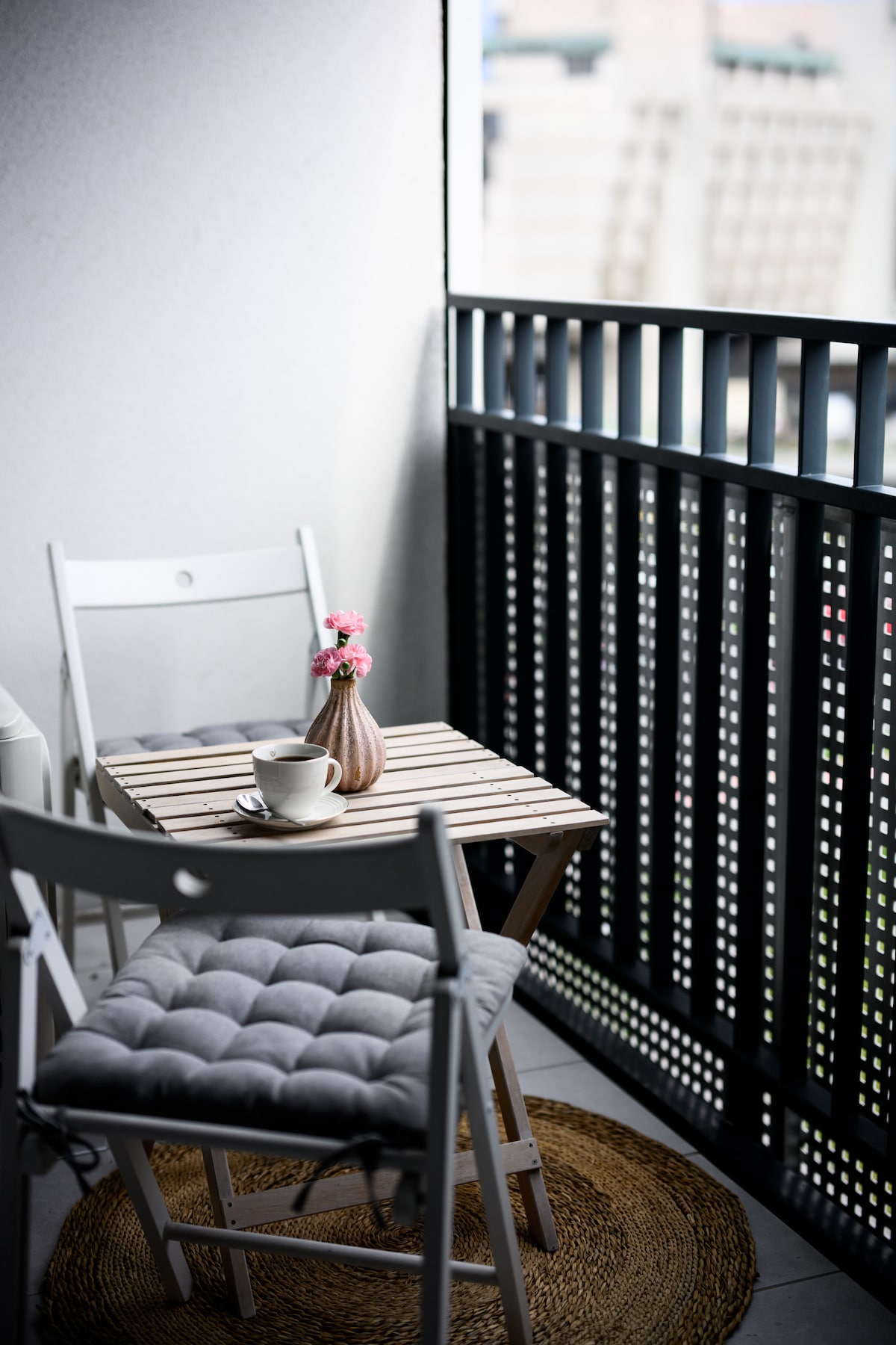 A small balcony area is presented with a wooden table and two white chairs. A vase with pink flowers adds a touch of color, while a cup rests on the table. A natural fiber rug enhances the space next to the chairs, with a view visible through the railing.