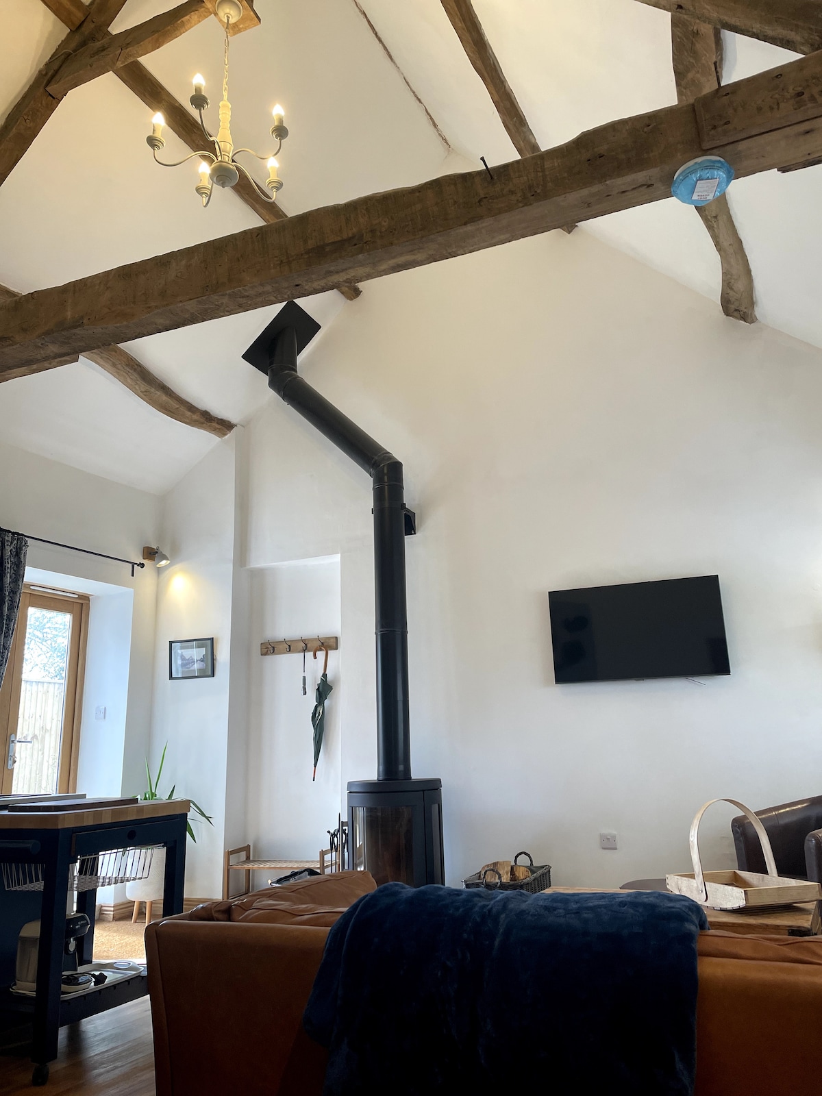 An open-plan living area features rustic wooden beams and a tall black flue leading to a stove. A flat-screen television is mounted on the wall, while a small table offers a space for dining. Natural light fills the room, highlighting the warm decor.