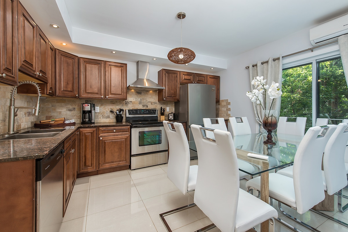 A modern kitchen features wooden cabinetry and stainless steel appliances, including an oven and refrigerator. A glass dining table with white chairs accommodates multiple guests. Natural light streams in through large sliding doors that open to a garden view, enhancing the spacious atmosphere.