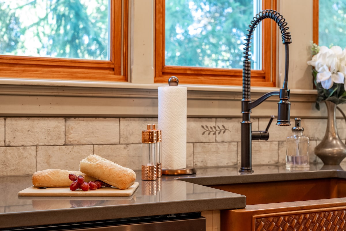 A modern kitchen countertop showcases a cutting board with two bread rolls and a cluster of grapes. A sleek faucet is positioned above a stainless steel sink, with decorative items, including a paper towel holder and a small glass container, visible on the counter.