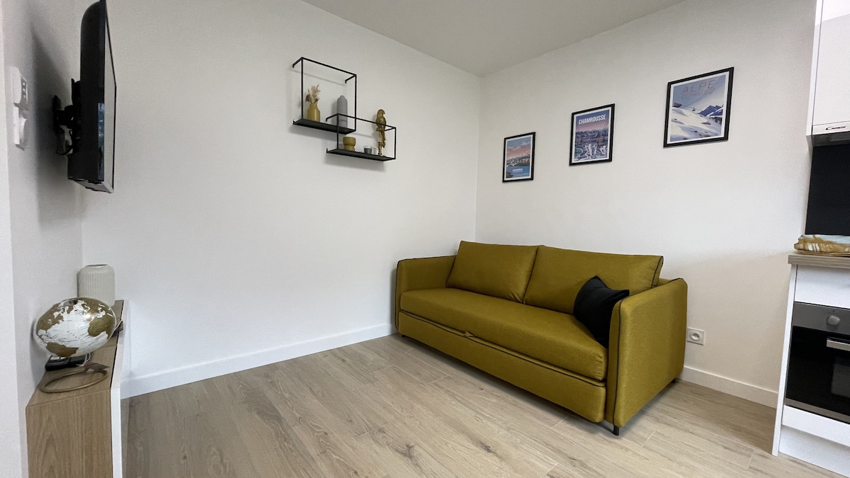 A cozy sitting area features a mustard yellow sofa against a white wall. Various framed photographs are arranged above the sofa. A minimalist shelf displays decorative items, while a television is mounted on the wall. Light wood flooring creates a warm ambiance.