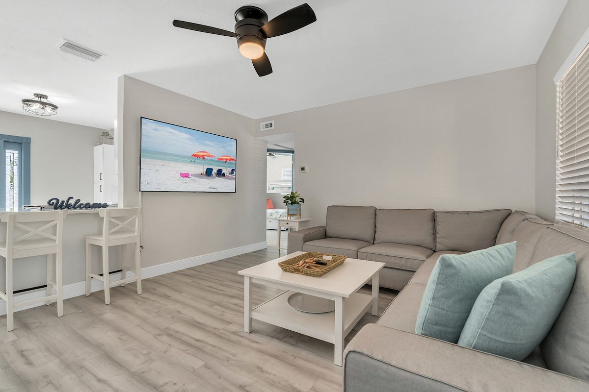 A comfortable living area is shown with a light-colored sectional sofa and a small coffee table. A wall-mounted TV is visible, displaying a beach scene. Natural light filters through the windows, enhancing the neutral tones of the decor.