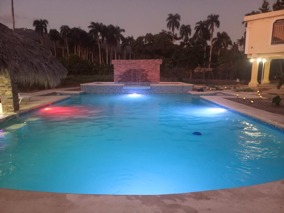 A spacious pool is illuminated with soft blue and red lights, creating a serene ambiance at dusk. Surrounding palm trees provide a lush backdrop, while a stone feature adds visual interest. The pool area includes a jacuzzi, enhancing the recreational setting.