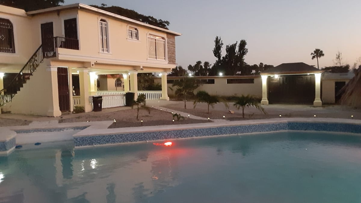 A two-story building is visible beside an inviting pool, with soft lights illuminating the surroundings. The pool features a gentle blue hue and is bordered by landscaped areas. The structure has multiple access points, and a peaceful evening ambiance is created as dusk settles.
