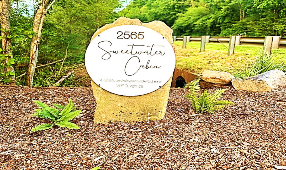 A stone marker displays the cabin name 'Sweetwater Cabin' and the address, set against a backdrop of lush green foliage and a gravel path. The inviting natural surroundings create a welcoming entrance for guests arriving at the property.