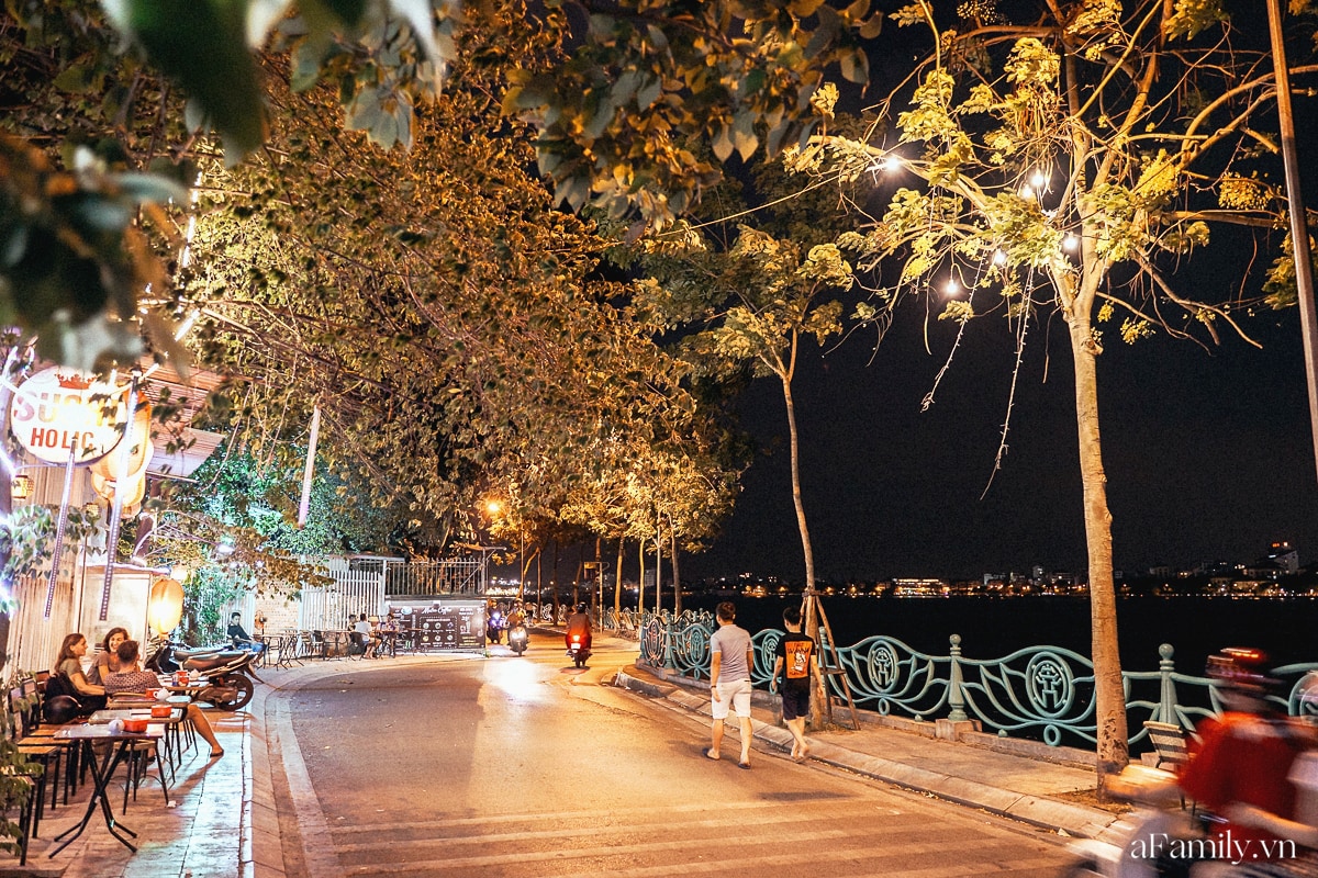 A street scene along West Lake is depicted at night, illuminated by soft lights hanging from trees. Individuals are seen walking along the sidewalk, with dining tables and chairs positioned nearby, creating a communal atmosphere by the lakeside.