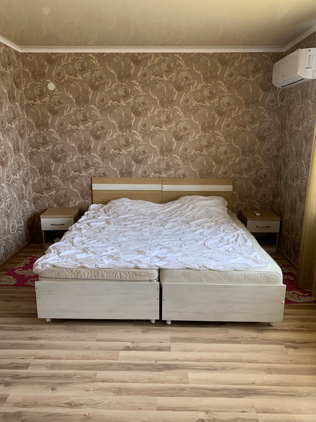 A bedroom features a spacious bed with a crumpled white blanket, placed against a patterned wall. Two nightstands are positioned on either side of the bed, complementing the warm wooden floor. Natural light streams in through a nearby window.