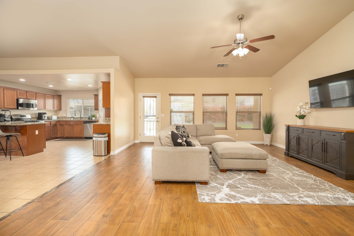 A spacious living area features a large sectional sofa positioned on a neutral-toned area rug. Natural light fills the room through several windows, highlighting the wooden flooring. A flat-screen TV is mounted on the wall, while the open layout connects to the kitchen in the background.