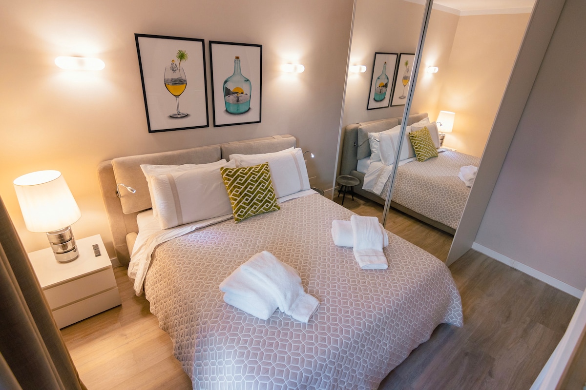 A cozy bedroom is presented with a spacious bed adorned with white linens and a patterned blanket. A colorful cushion adds a pop of color. Framed artwork is displayed above the bed, and soft lighting is provided by two mounted sconces. A mirrored closet expands the room's visual space.