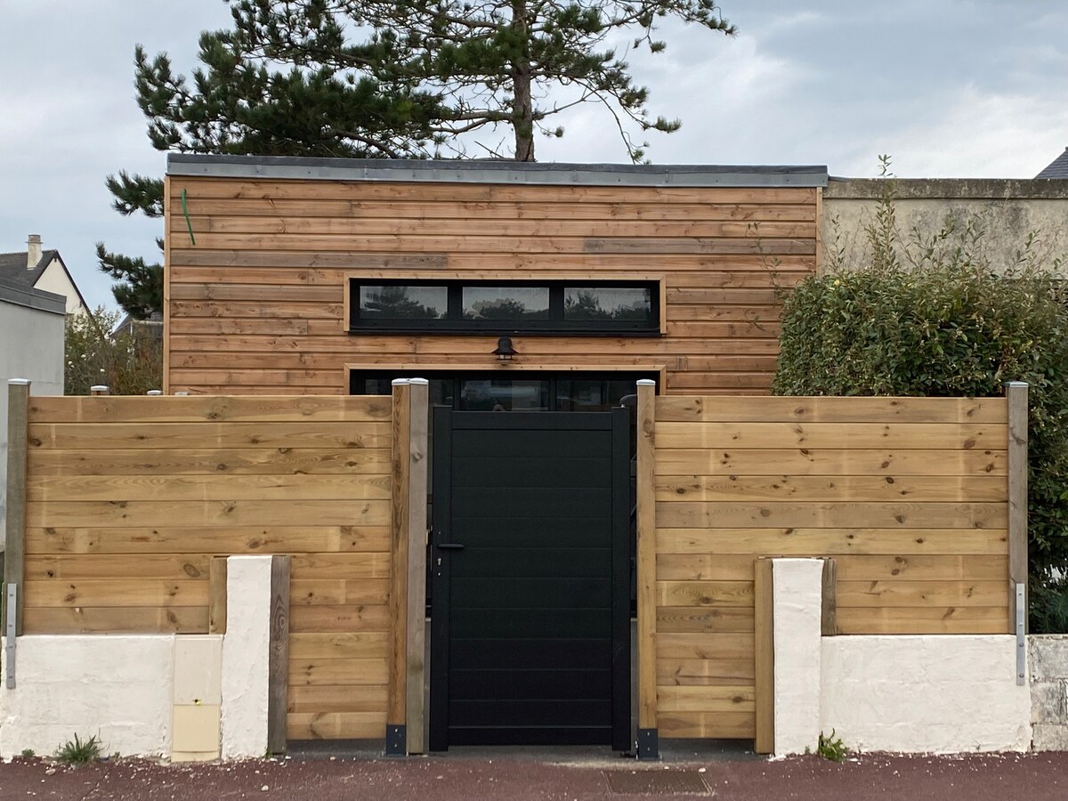 The exterior of the property features a modern wooden facade, framed by tall wooden fencing. A black gate provides access, further enhancing the privacy of the space. Large windows are visible at the top, offering a glimpse of natural light within.