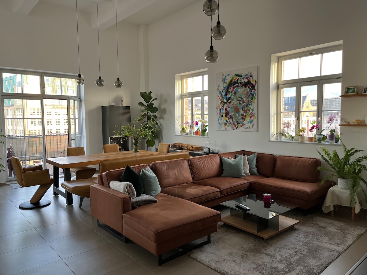 A modern living space features a spacious brown sectional sofa with decorative pillows, a glass coffee table, and a wooden dining table surrounded by chairs. Tall windows allow natural light to flood the room, while potted plants add a touch of greenery to the decor.