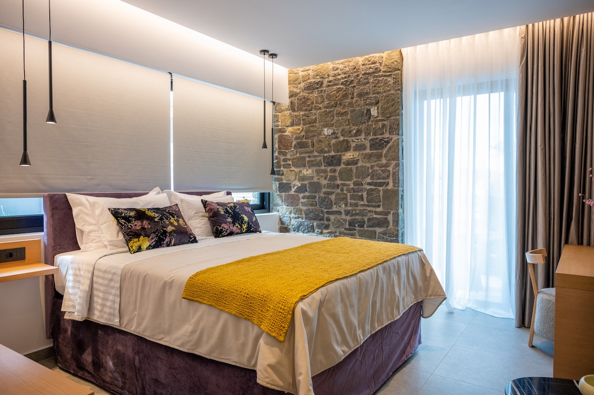 A spacious sleeping area features a large bed with a plush headboard and decorative pillows. The room is enhanced by stone accent walls and natural light filtering through sheer curtains. A cozy yellow throw is draped over the bed, complementing the modern design.