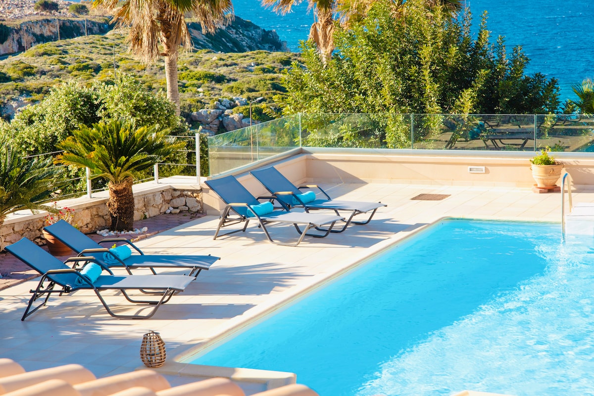 A large pool is surrounded by several lounge chairs, inviting relaxation. Lush greenery and palm trees are visible in the background, complemented by a clear view of the sea. Clear glass panels provide safety and unobstructed views of the serene surroundings.