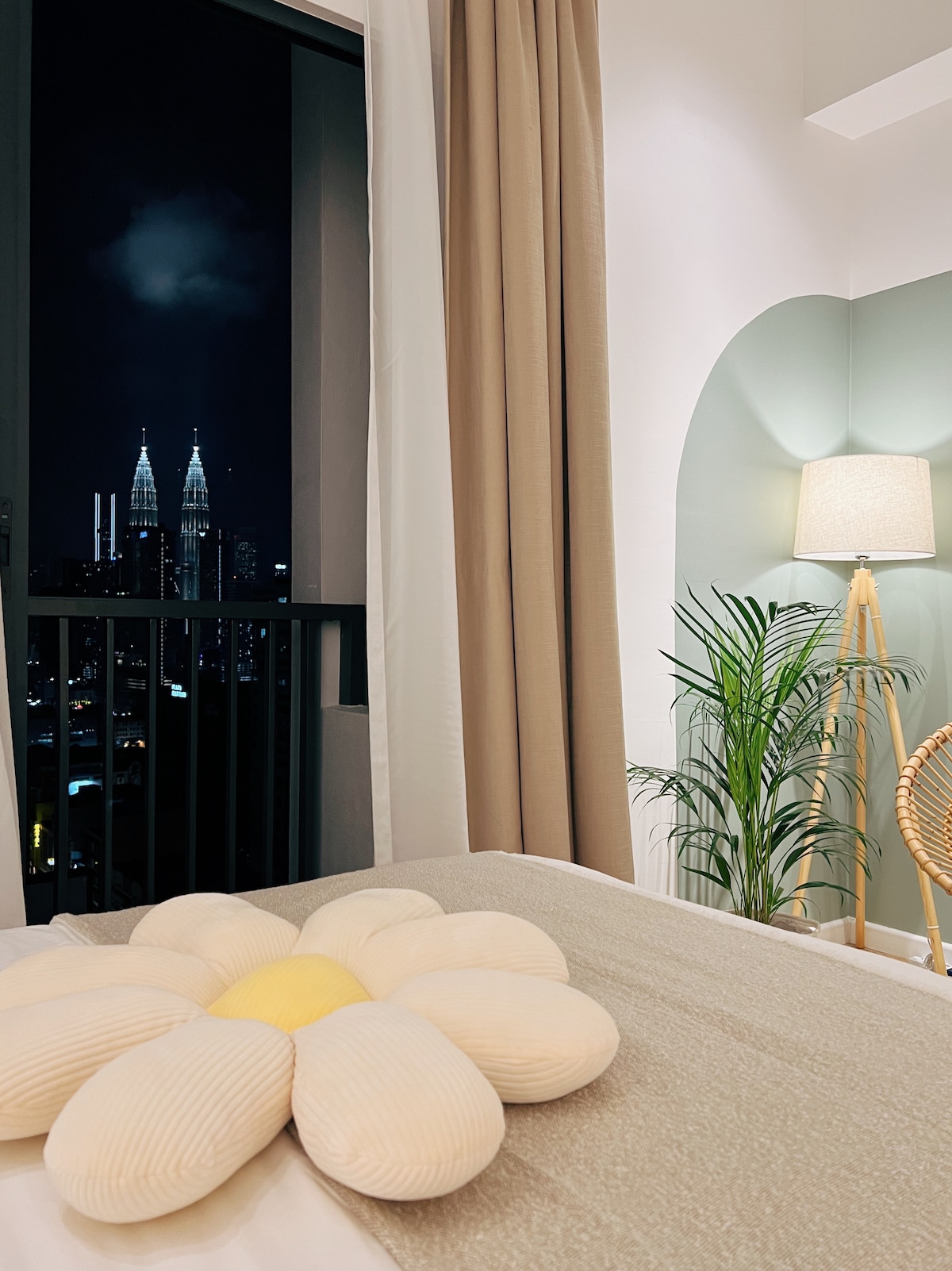 A cozy studio space showcases a queen bed adorned with a plush flower-shaped pillow. The nearby window reveals a view of the illuminated KLCC Twin Towers against the night sky. Soft lighting from a standing lamp complements the warm color palette of the room.