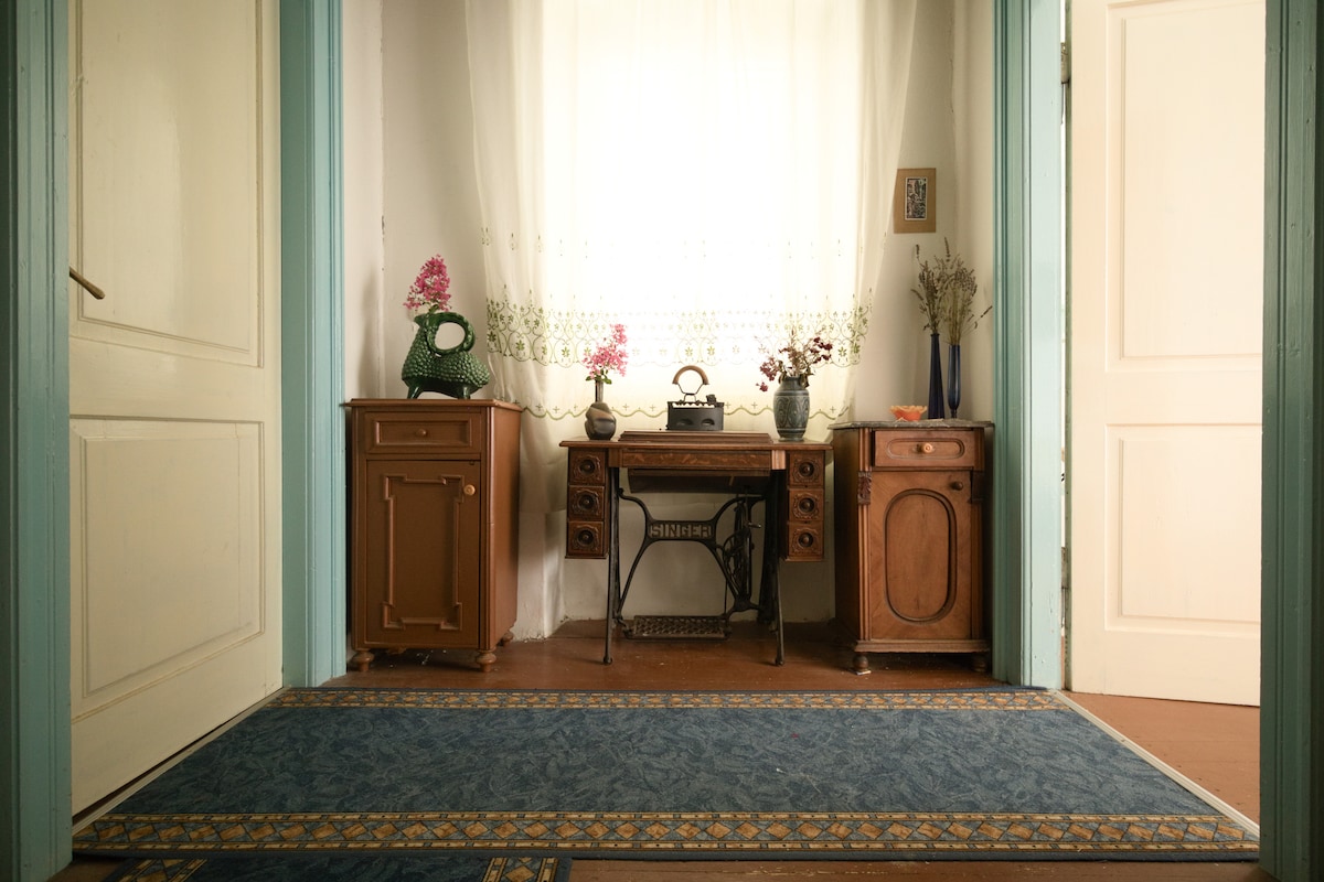 A welcoming entryway features a blue patterned rug and vintage furnishings. A small desk is positioned against the wall, complemented by decorative flowers in vases. Natural light filters through a sheer curtain, creating a soft ambiance that highlights the elegant wooden cabinetry.