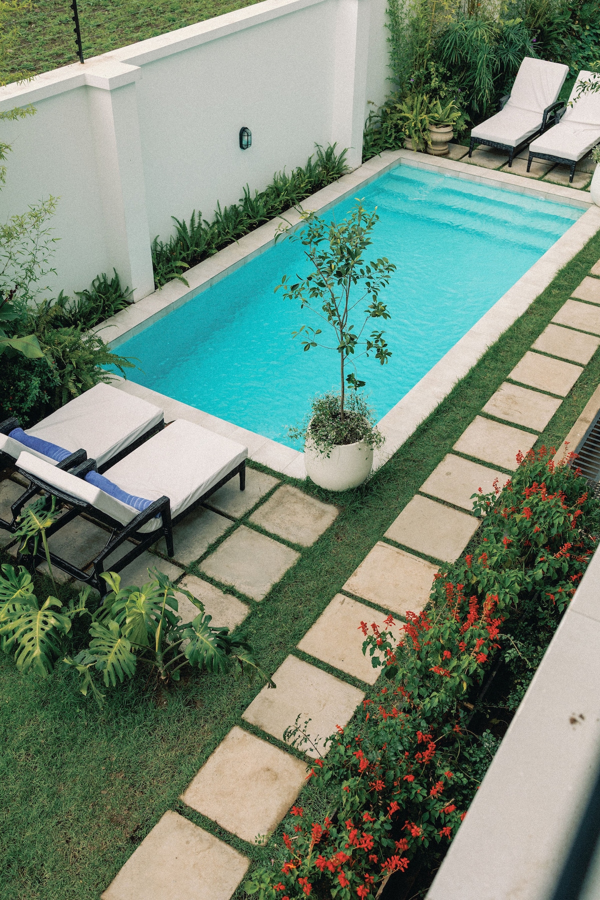 A serene pool area is framed by a well-maintained garden, featuring a clear blue pool bordered by decorative stones and lush greenery. Two lounge chairs are positioned beside the pool, inviting relaxation under natural light. Colorful flowers add vibrancy to the landscape.