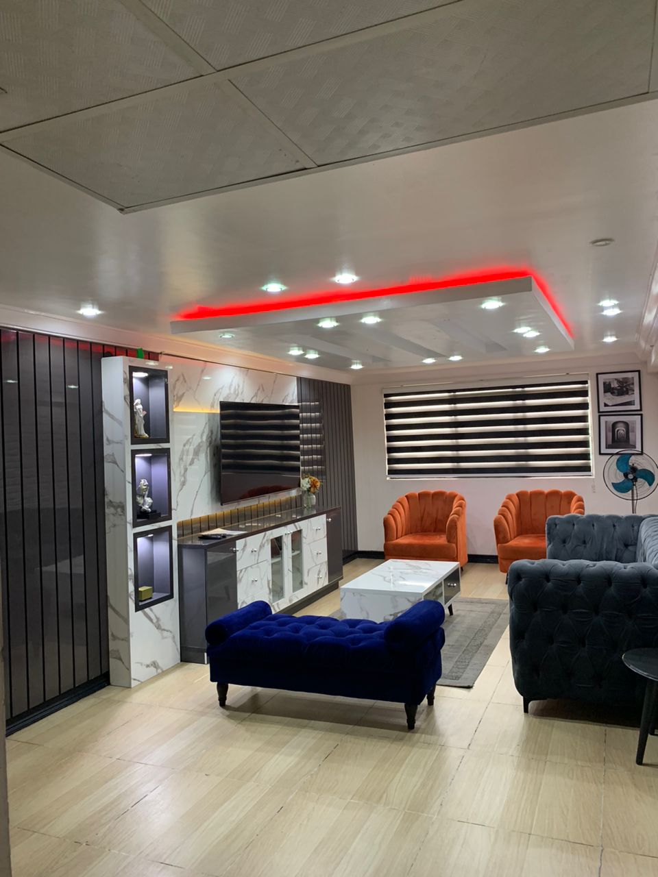 A modern living area is presented with a combination of stylish furniture. Two orange armchairs and a navy blue sofa provide comfortable seating. A sleek coffee table sits in the center, complemented by a wall-mounted TV and decorative lighting along the wall.