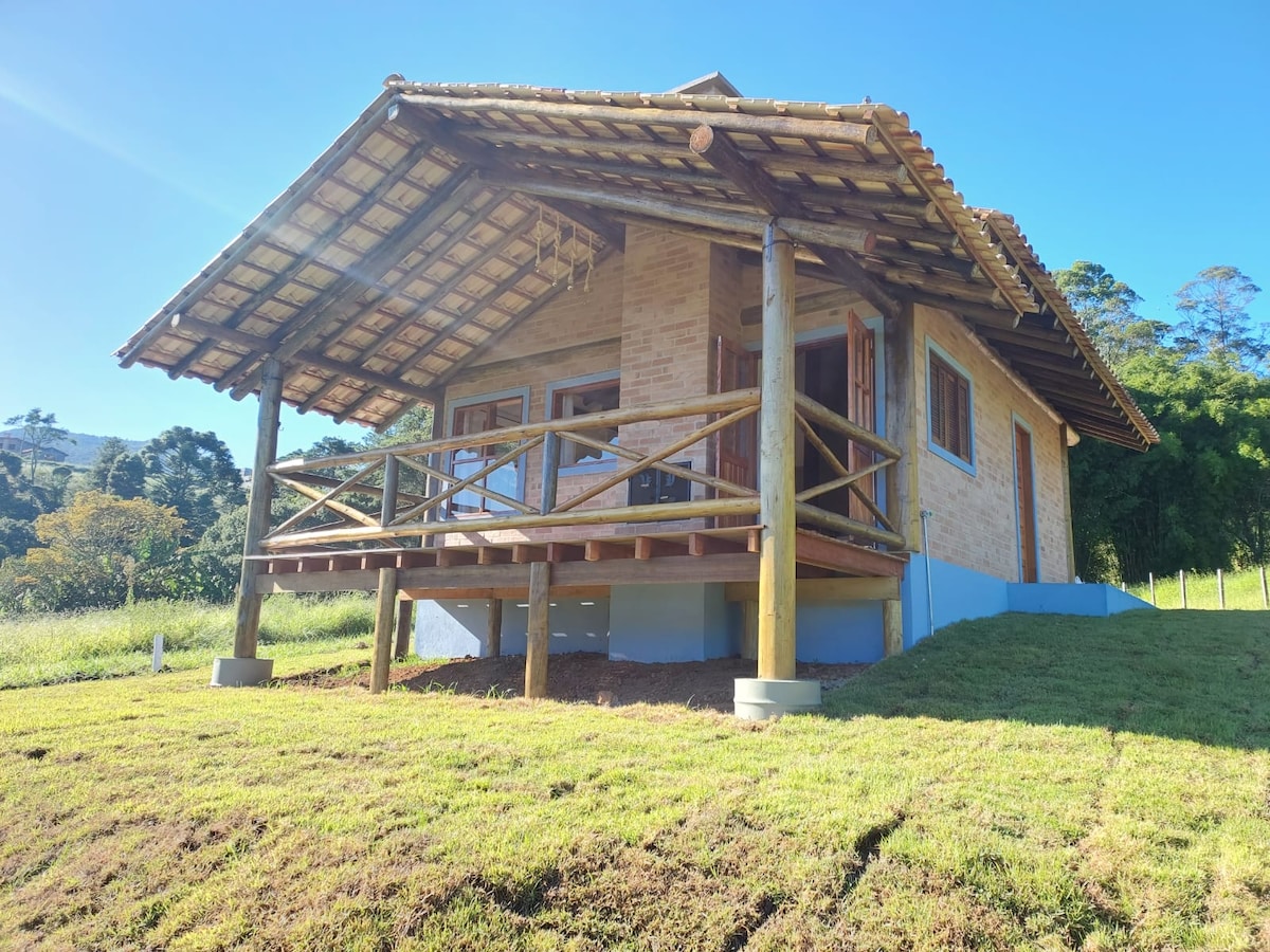 A rustic chalet features a spacious front porch supported by logs, two wooden rocking chairs are arranged for relaxation. The exterior is crafted from earthy tones of brick, surrounded by green grass and trees, showcasing a peaceful integration with nature.