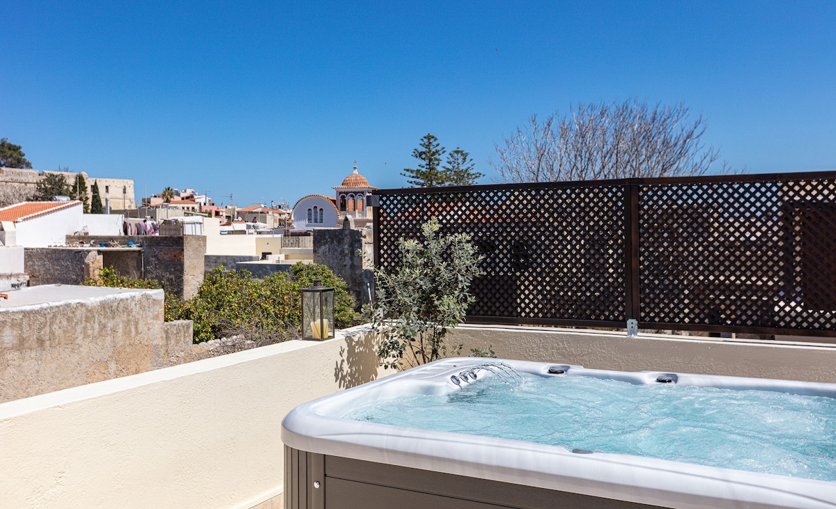 A jacuzzi is positioned on a spacious terrace, providing a view of the surrounding rooftops and the distant Venetian Castle. Sunlight reflects off the water, enhancing the inviting atmosphere of relaxation. Greenery is visibly present nearby, adding a natural element to the setting.