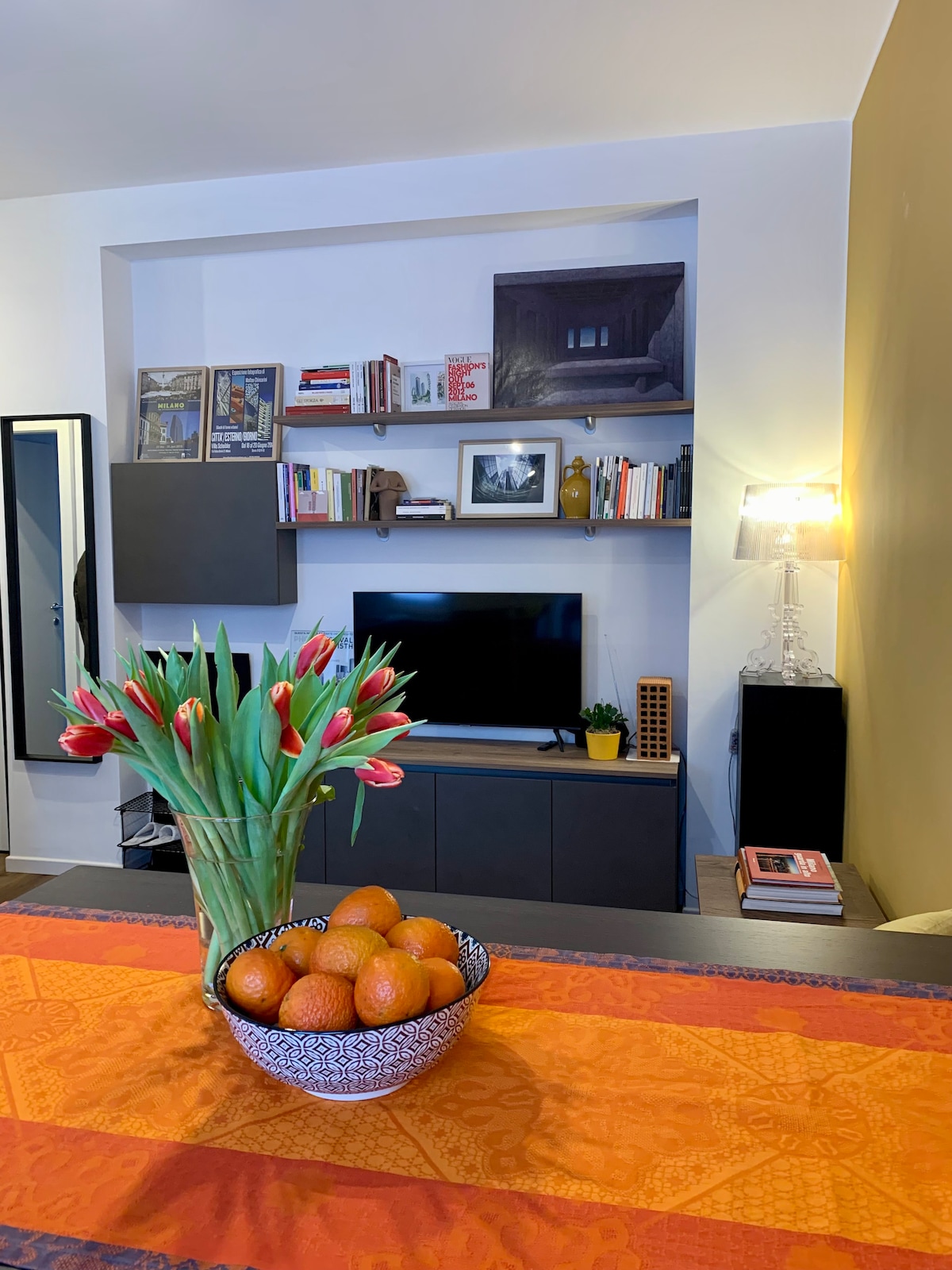 A decorative arrangement features a bowl of oranges and a bouquet of tulips on a table. In the background, a modern shelving unit displays books and artwork, while a flat-screen television is positioned atop it. The room is illuminated by a bedside lamp.