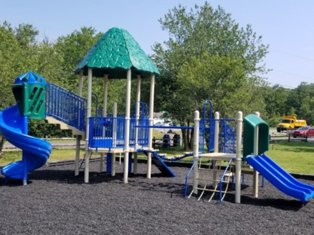 A colorful playground structure is visible, featuring multiple slides and climbing areas. The bright blue and green equipment contrasts with the dark rubber surface beneath. Surrounding trees provide a natural backdrop, enhancing the outdoor play environment.