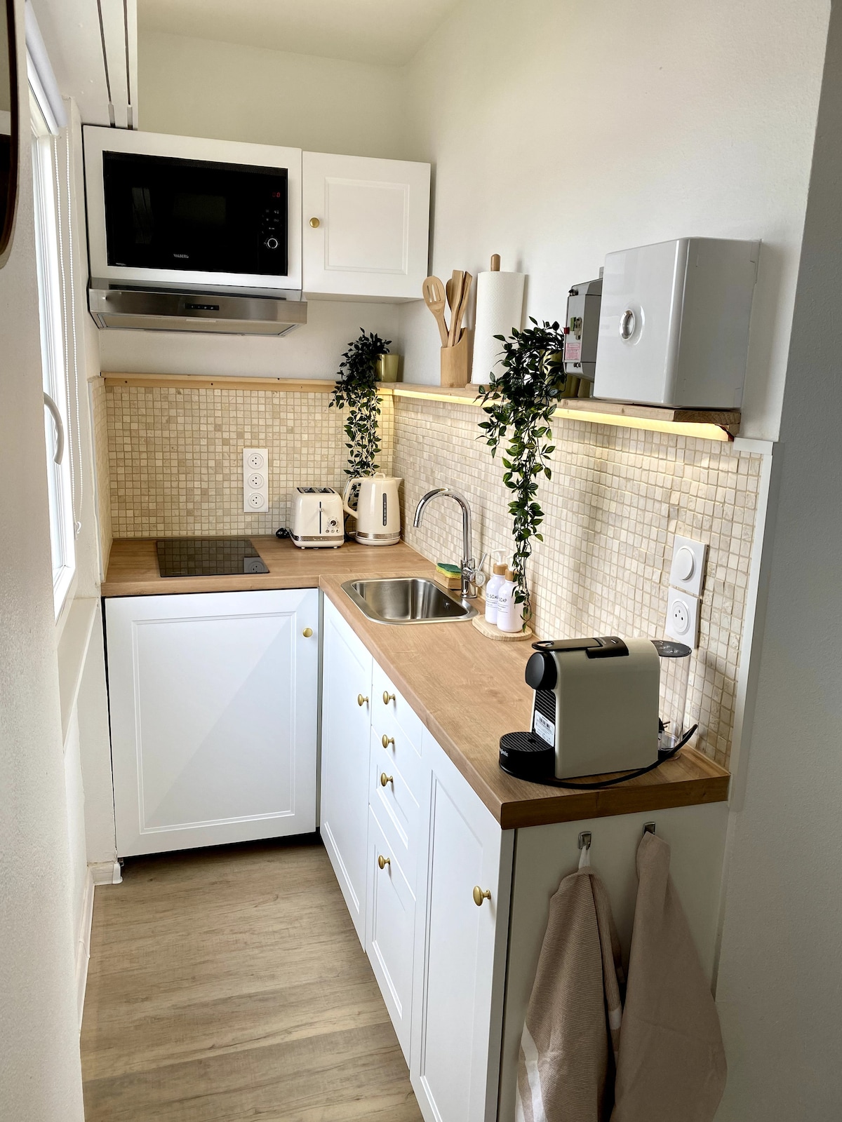 A compact kitchen features white cabinetry and a wooden countertop. Essential appliances, including a coffee machine, kettle, and microwave, are neatly arranged. A tile backsplash adds texture, while under-cabinet lighting enhances visibility. Small plants provide a touch of greenery, contributing to a functional cooking space.