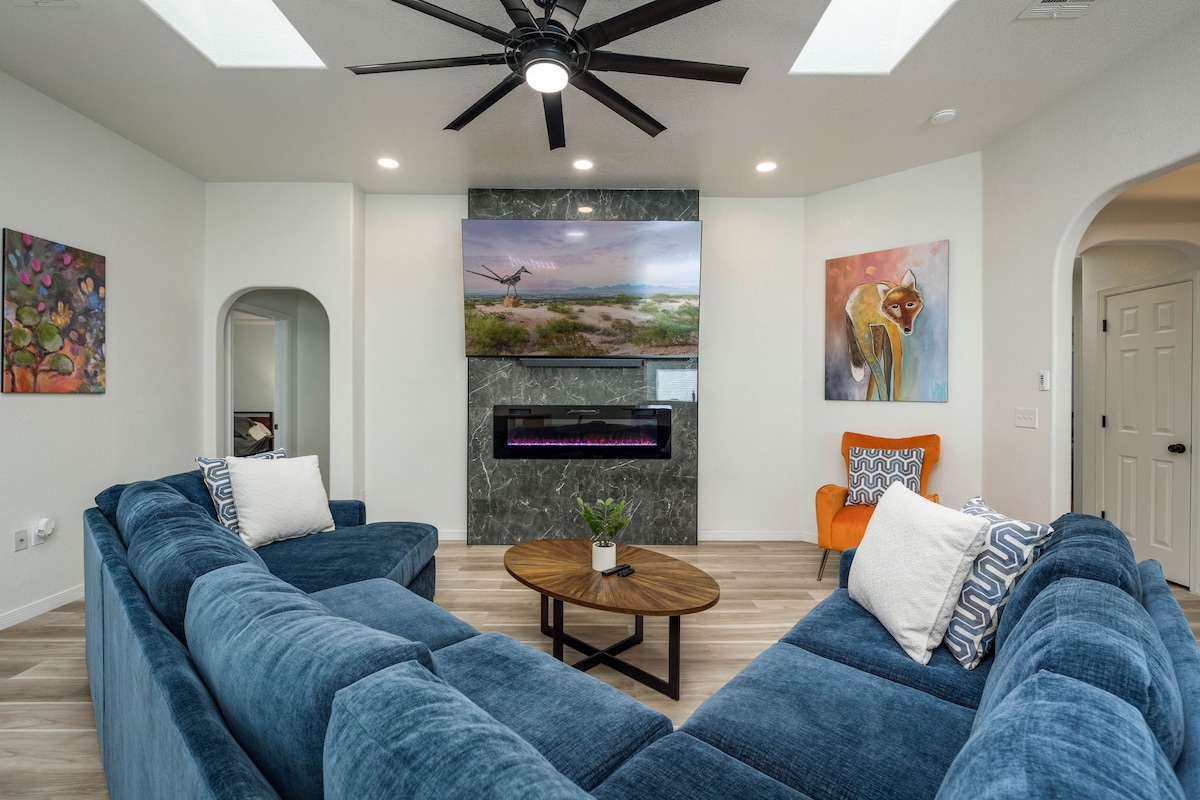 A spacious living room features a large blue sectional sofa arranged around a circular coffee table. A mounted flat-screen TV is above a modern electric fireplace. Colorful artwork adorns the walls, and an orange accent chair adds a pop of color. Light is provided by a ceiling fan and natural light from nearby openings.