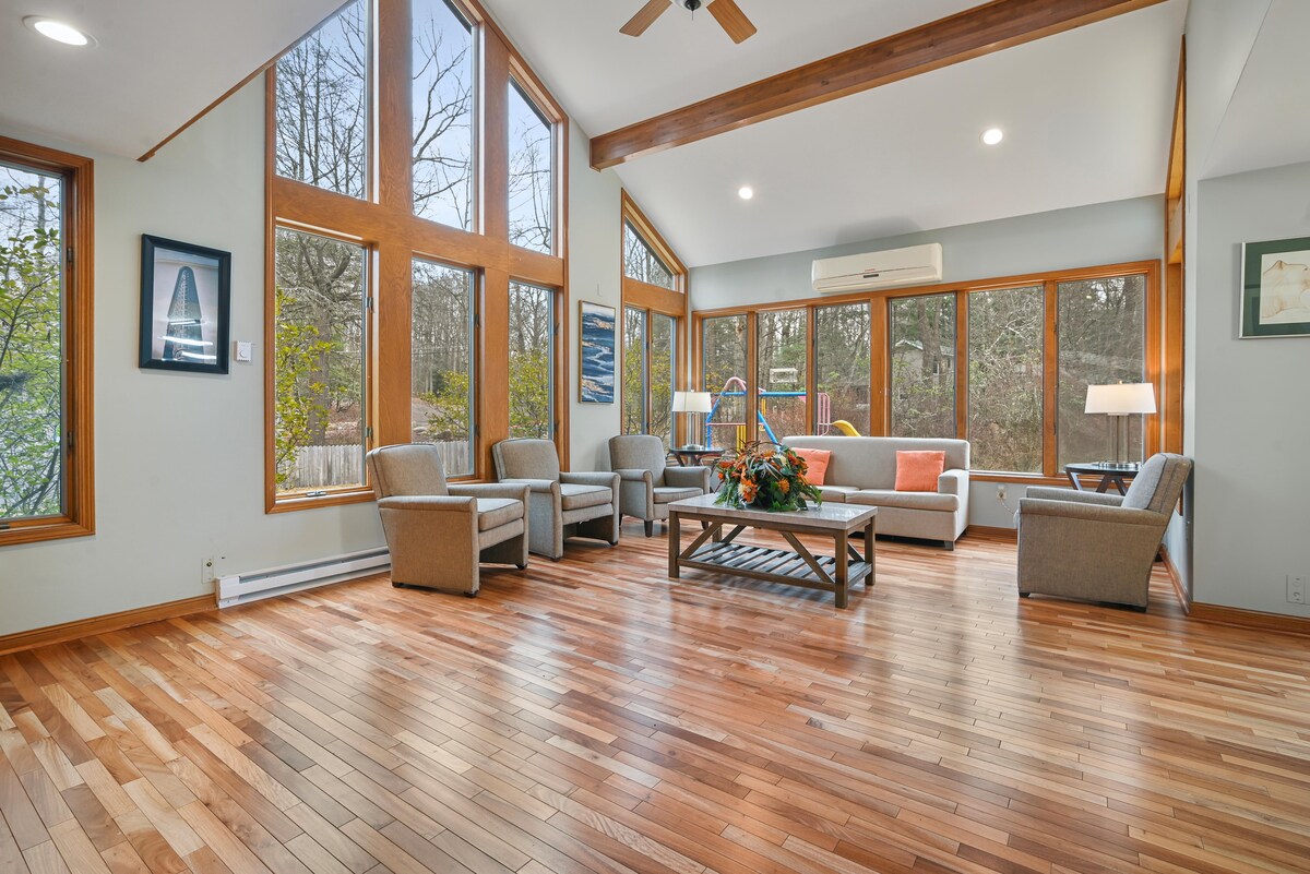 A spacious living area features large windows that allow natural light to fill the room. Comfortable seating arrangements, including multiple chairs and a sofa, are positioned around a central coffee table. The hardwood floor adds warmth to the inviting space.