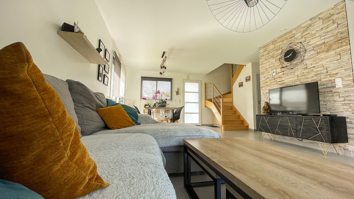 A spacious living area is presented, featuring a modern sofa adorned with colorful cushions. A wooden coffee table is positioned in front, while a flat-screen television is placed on a sleek console. Natural light fills the room, highlighting the stylish decor and elegant staircase.
