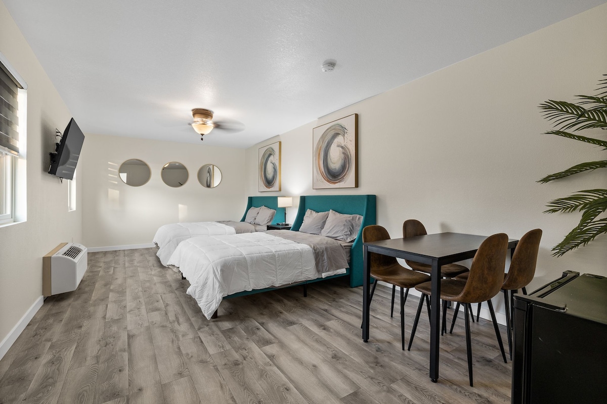 A well-lit room features two spacious queen beds with light bedding, complemented by decorative wall art. A dining table with four chairs occupies one corner, while mirrors and a television are mounted on the wall, enhancing the sense of space and comfort.