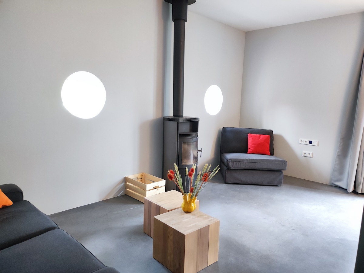 The cozy living area features a wood stove, enhancing warmth. Neutral walls are complemented by a sofa with vibrant cushions. Two wooden side tables are positioned nearby, and sunlight enters through circular windows. A decorative vase with fresh flowers adds a pop of color.