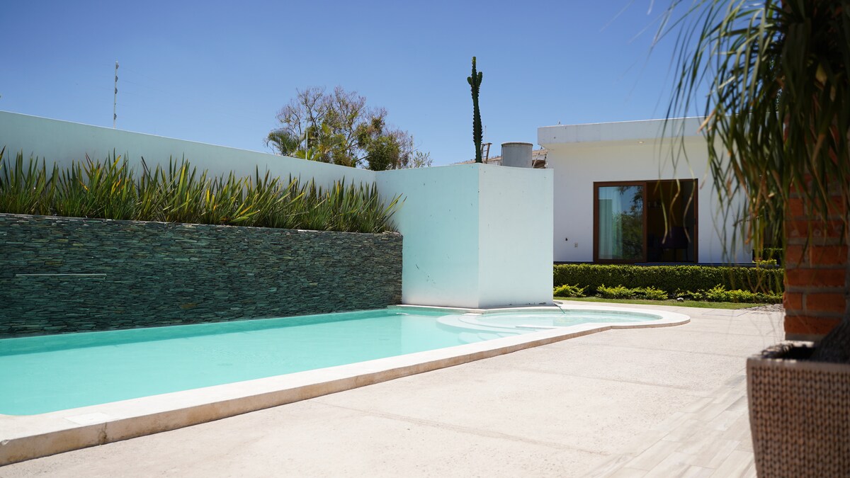 A serene outdoor space is highlighted by a well-maintained swimming pool, featuring clear turquoise water. The area is surrounded by lush green plants and a stone wall, providing a relaxing environment. A house is visible in the background, framed by clear skies.