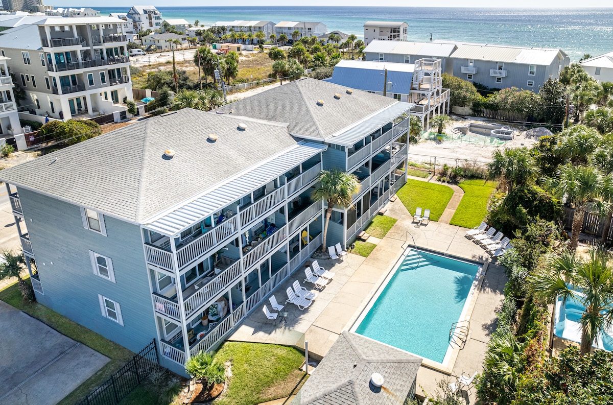 An aerial view showcases a three-story building with a light blue exterior, surrounded by lush greenery and a sparkling pool area. Loungers are placed around the pool, providing a relaxing outdoor space. The coastline and ocean are visible in the distance, contributing to a serene atmosphere.