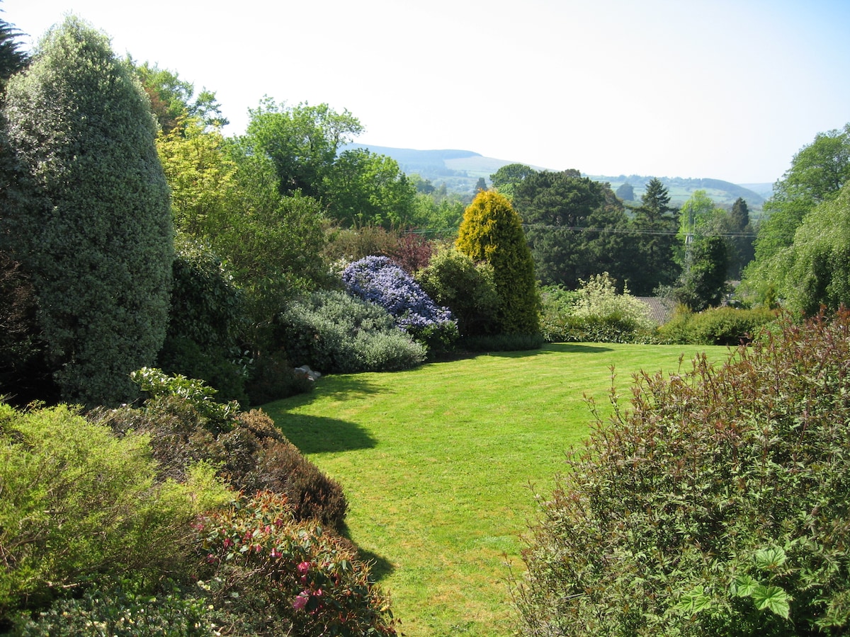 A lush garden is showcased, featuring a variety of trees and flowering shrubs. A well-maintained green lawn occupies the center, bordered by vibrant foliage. The background reveals distant hills under a clear blue sky, contributing to the serene atmosphere of the outdoor space.