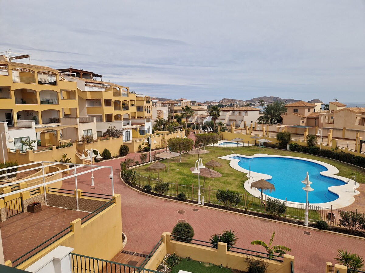 The view from the terrace captures a landscaped area with a large swimming pool surrounded by green lawns. Sun loungers and umbrellas are positioned near the pool. The residential buildings feature multiple levels, creating a communal atmosphere. The sky is mostly overcast.