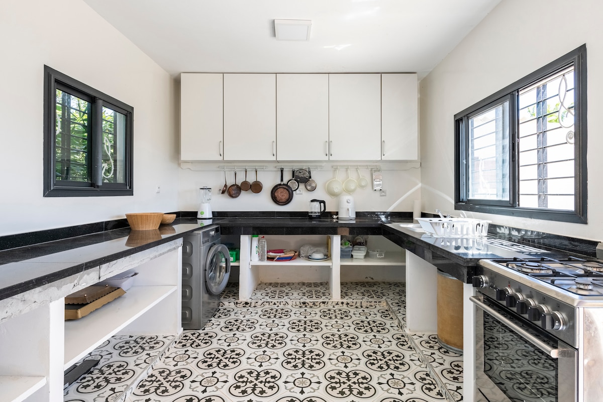 A well-equipped kitchen is presented with modern appliances and ample storage. Dark countertops, white cabinetry, and a decorative tiled floor create a functional cooking space. Large windows allow natural light to brighten the area, showcasing a combination of culinary tools displayed on the wall.