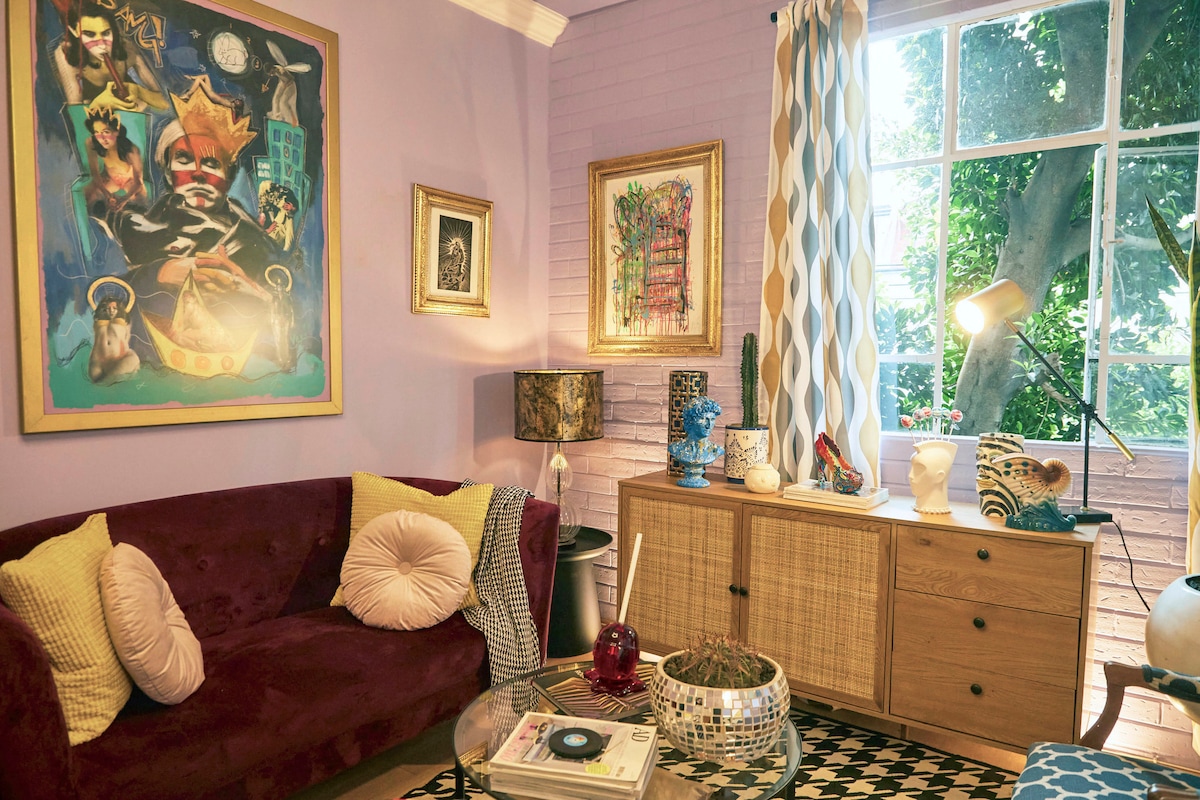 A cozy living area is highlighted by a deep red sofa adorned with decorative pillows. A vibrant piece of wall art attracts attention, complemented by natural light flowing through large windows. A wicker cabinet and a variety of decor elements add to the inviting environment.