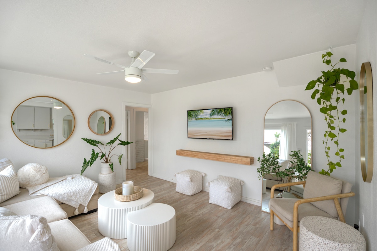 A bright living room features white walls and light wooden flooring. A comfortable seating area is arranged with various textured cushions, complemented by round mirrors on the wall. A television is mounted above a wooden shelf, while green plants add vibrancy to the space.