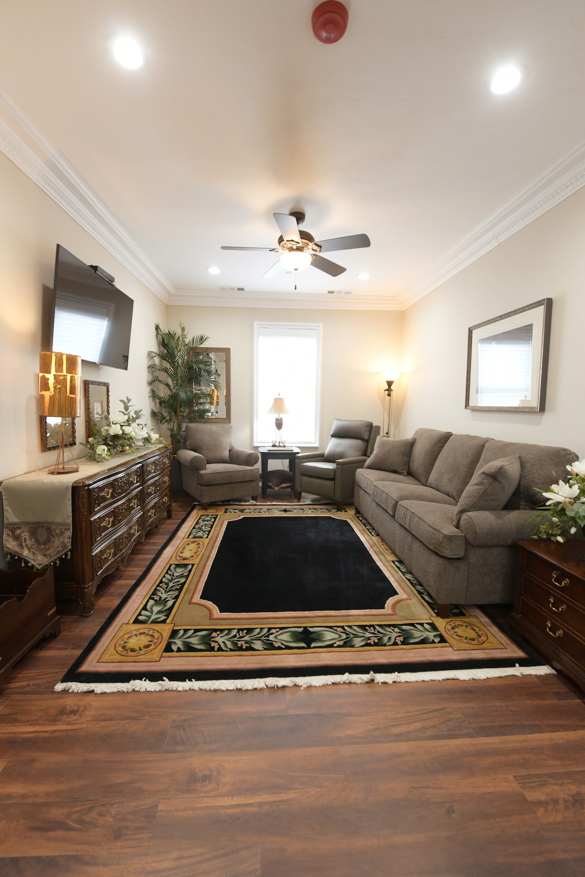 A comfortably furnished living room is presented with a soft gray sofa and recliner chairs. A dark area rug is placed on polished wood flooring. A decorative console table and a television accompany elegant lighting near a window that allows natural light to fill the space.