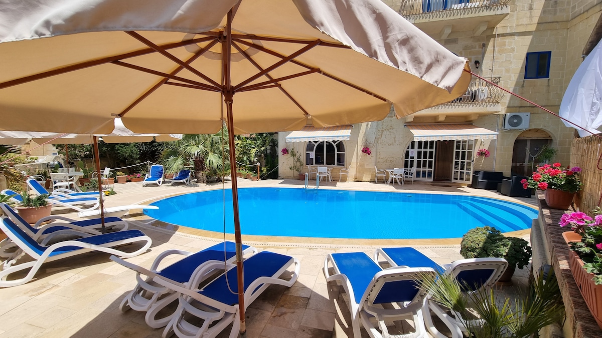 An inviting pool area is surrounded by lounge chairs and large umbrellas, providing shade. Potted plants and flowers add greenery to the scene, while a spacious terrace and glass doors are visible in the background, enhancing the outdoor relaxation space.