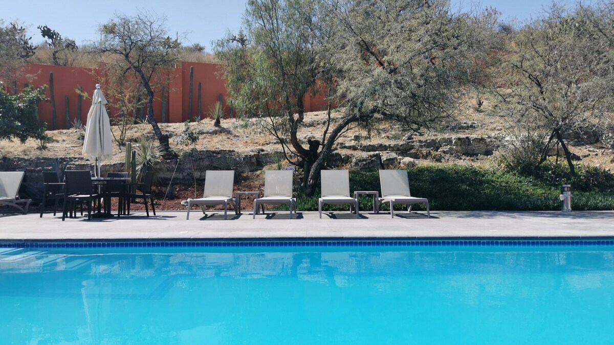 A clear pool reflects the sky, surrounded by sun loungers positioned on a spacious deck. Lush greenery and sparse trees create a natural backdrop, with a shaded area featuring an umbrella and a table set nearby.