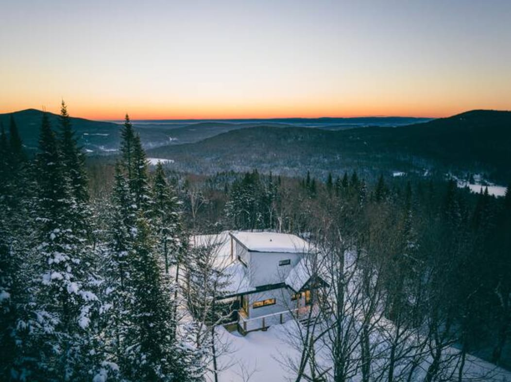 The cabin is situated amidst a snowy landscape, surrounded by tall evergreen trees. A breathtaking sunset casts warm colors across the horizon, contrasting the cool tones of the winter scene. The mountainous terrain is visible in the distance, enhancing the serene natural setting.