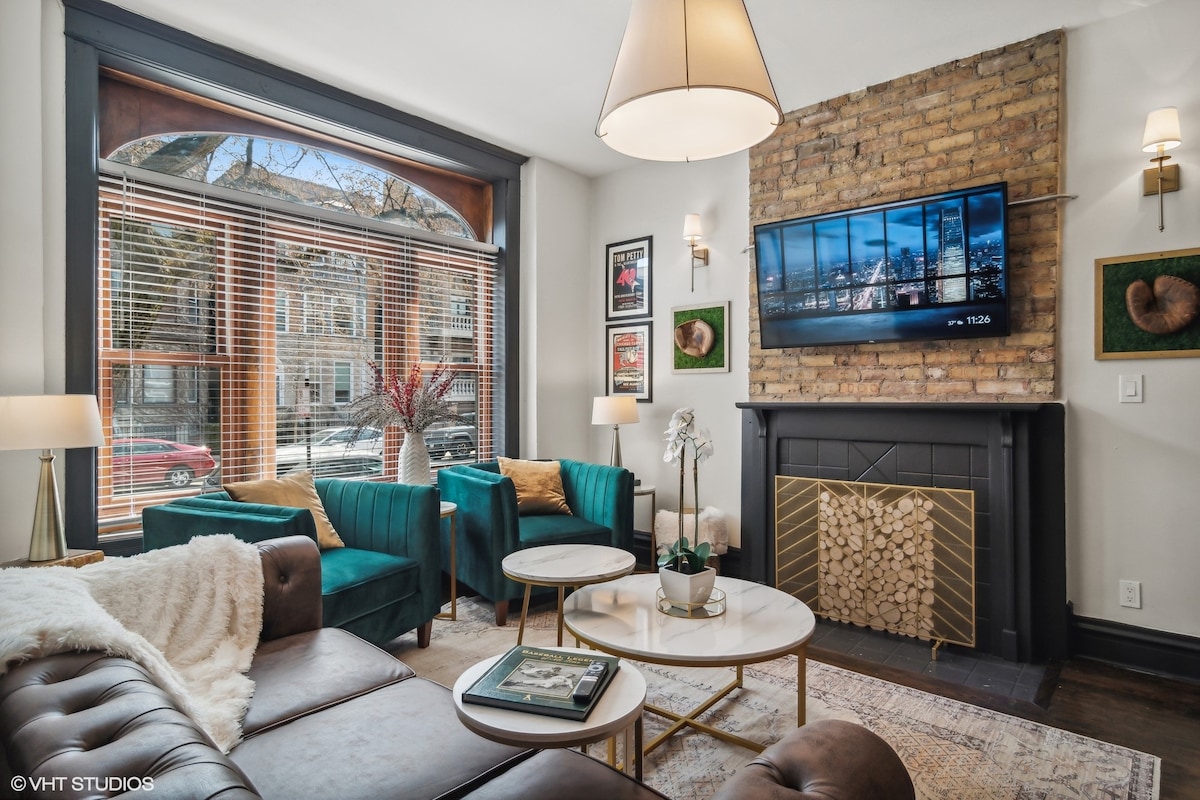A cozy living area is shown with large windows allowing natural light to fill the space. Two green armchairs and a brown leather sofa are arranged around two circular coffee tables. A mounted flat-screen TV is positioned above a decorative fireplace, accompanied by exposed brick walls.