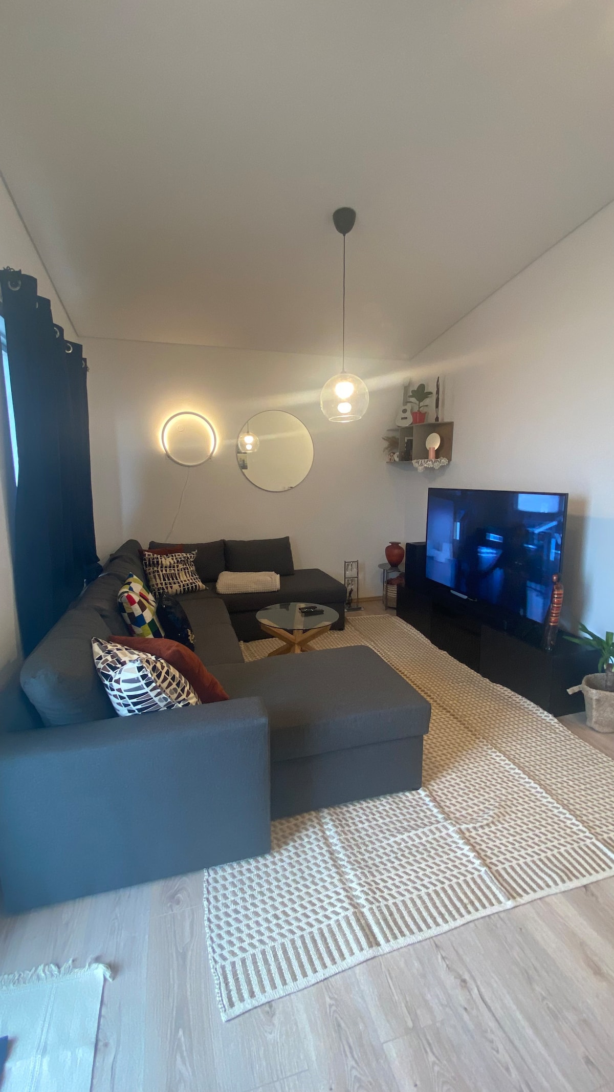 A comfortable living area features a sectional sofa adorned with colorful cushions. A large television is mounted on the wall opposite the seating, with a round mirror and ambient lighting enhancing the space. A textured area rug covers the floor, adding warmth.