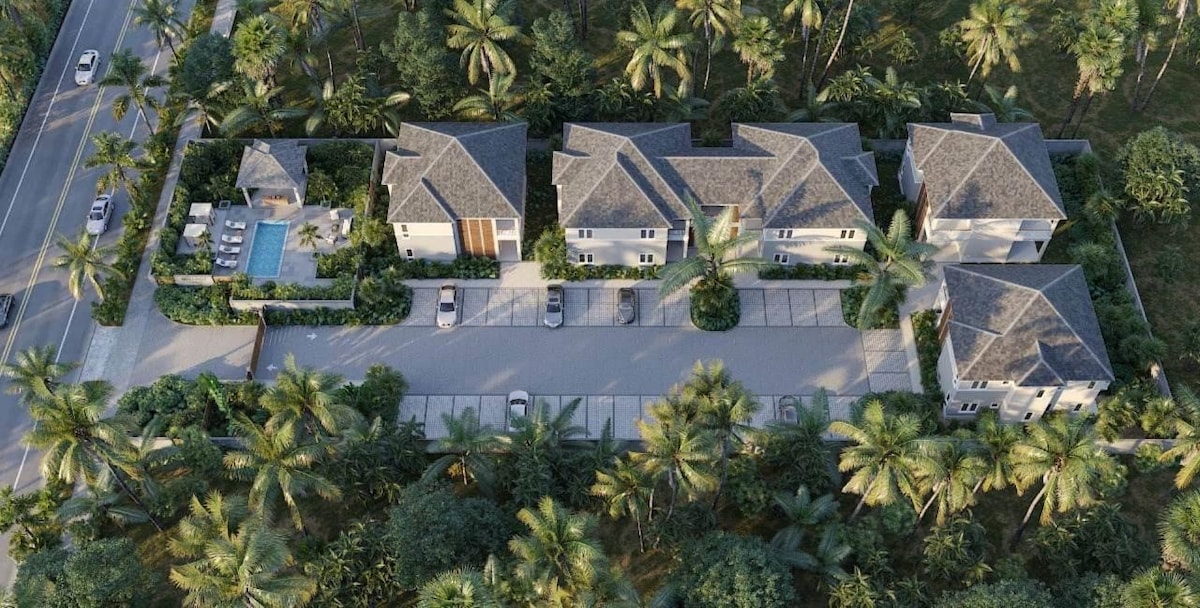 An aerial view reveals a gated boutique development surrounded by lush greenery and towering palm trees. The complex features several buildings arranged along a central roadway, with a pool area and parking spaces visible, showcasing a tranquil and inviting atmosphere.