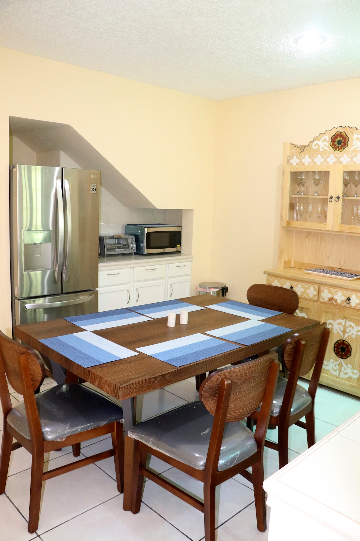 A well-appointed kitchen features a wooden dining table with four chairs, accompanied by a decorative hutch. Modern appliances, including a refrigerator and microwave, are visible. Light reflects off the tiled floor, contributing to a bright and functional space.