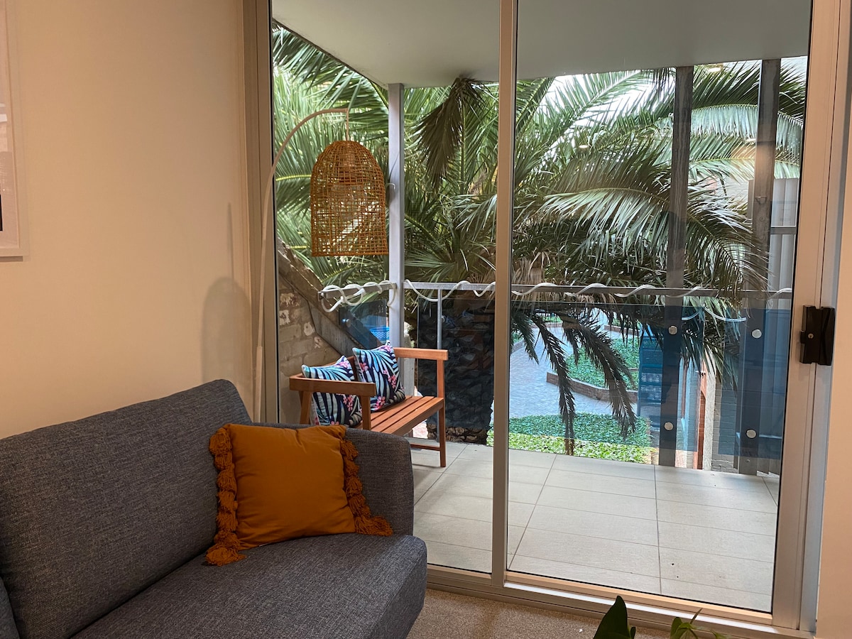 A cozy living area features a blue sofa adorned with a textured mustard pillow. Large glass doors open to a balcony surrounded by lush palm trees, bringing natural light into the space. A small wooden chair is positioned nearby, enhancing the serene atmosphere.