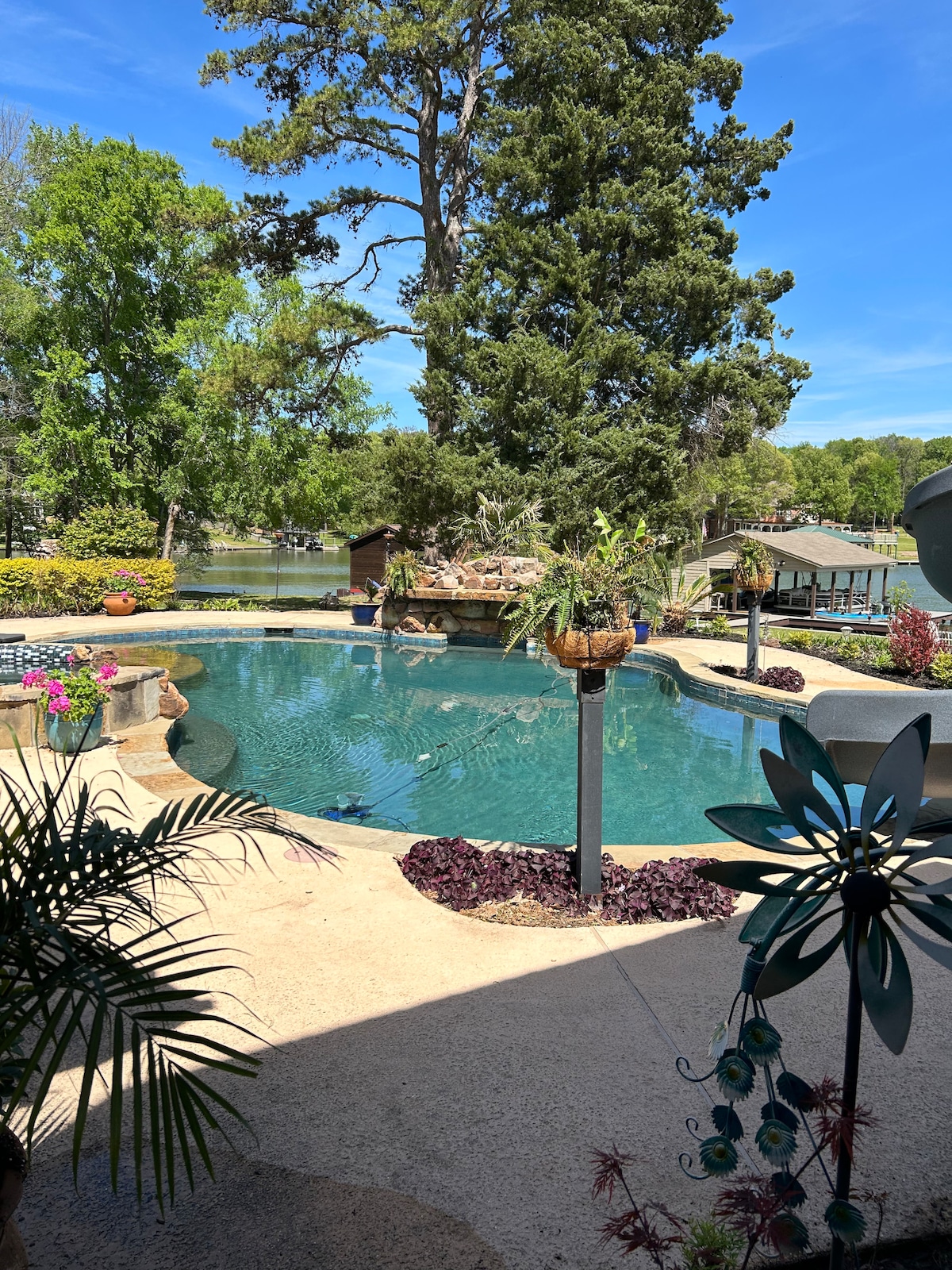 A serene swimming pool is surrounded by lush greenery and colorful flowers. The water reflects the sky, while a large tree provides shade nearby. In the background, a boathouse is visible along the water's edge, enhancing the peaceful atmosphere.
